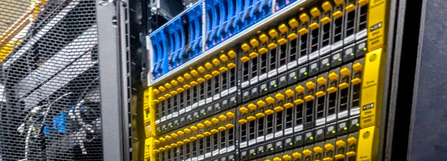 Primer plano de una cabina de almacenamiento en un rack de centro de datos, con filas de bahías de discos, pestillos amarillos y luces de estado verdes.