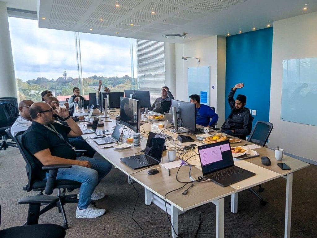 Bootcamp participants around a large table with laptops and monitors in a bright meeting room with a view over the city and trees.