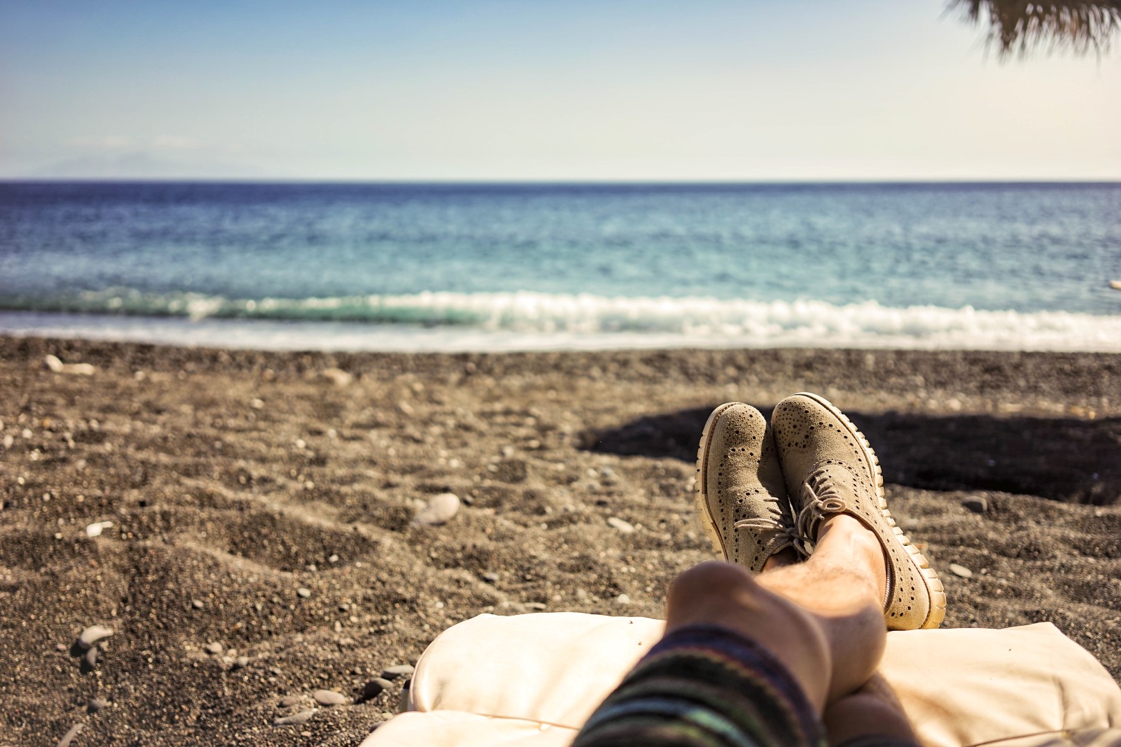 Man laying down on a beach chair relaxing looking at the sea. On the bottom left, the man's feet illuminated by the sun. Behind, blurred, some small waves breaking on the beach shore.